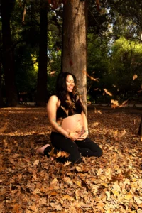 Mujer embarazada con conjunto negro sentada entre hojas de otoño durante una sesión de fotos de maternidad al aire libre.